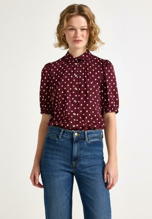 Ung kvinde iført en vinrød bluse med korte ærmer og hvide polkaprikker samt blå jeans med høj talje, stående foran en ensfarvet baggrund.