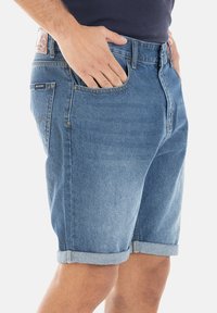 Shorts en denim, bleus, délavage moyen, ourlet retourné, cinq poches, patch en cuir avec logo au dos de la taille, fermeture à un bouton.