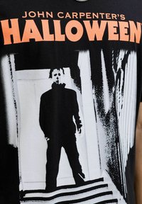 Crna pamučna majica s jakim narančastim natpisom "Halloween Johna Carpentera" iznad bijele grafike figure u mračnom okruženju.