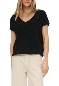 Žena oblečená v černém krátkém rukávu s výstřihem do V s jemnými texturovanými pruhy a světle béžových kalhotách, stojící v uvolněné poloze.