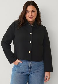 Black cropped jacket with a pointed collar, gold button closures, and side pockets, made from textured fabric. Worn over blue jeans.