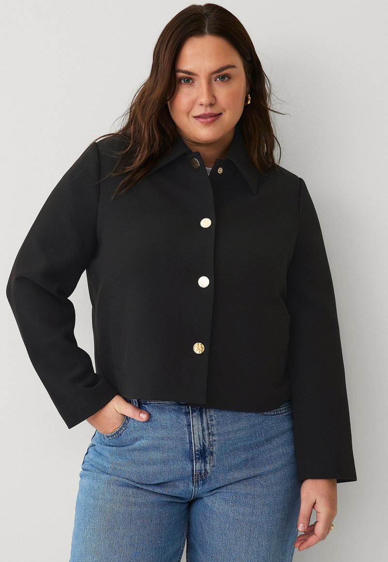 Black cropped jacket with a pointed collar, gold button closures, and side pockets, made from textured fabric. Worn over blue jeans.