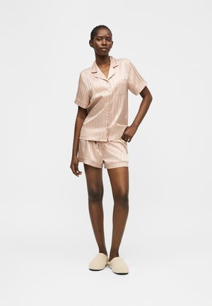 Sieviete stāv, valkājot bēša krāsas satīna īsas piedurknes pogātu kreklu un atbilstošas biksītes ar gaiši bēšām čībām vienkrāsainā fonā.