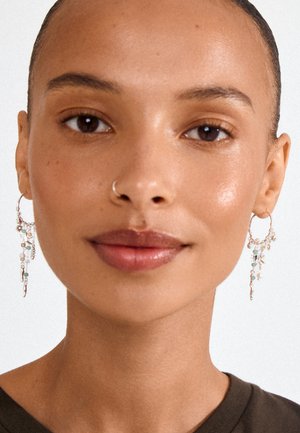 Noor naine, kellel on sile nahk ja tagasikeeratud juuksed, kannab hõbedasi rõngakujulisi kõrvarõngaid ripatsitega ja ninariiuliga.