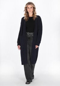 Marineblauer langer Cardigan aus einem weichen, fuzzy Material, mit offenem Frontbereich und zwei vorderen Taschen. Über einem schwarzen Oberteil und Jeans getragen.