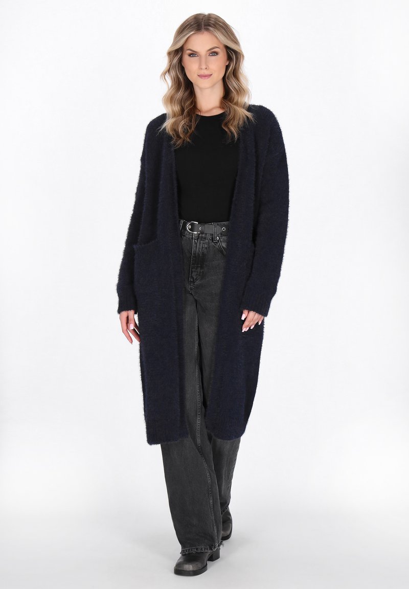 Cardigan lungo blu navy realizzato in un materiale morbido e peloso, con apertura frontale e due tasche anteriori. Indossato sopra una maglietta nera e jeans.