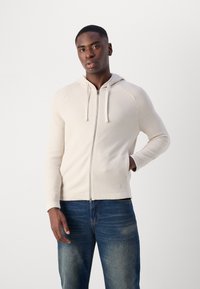 Lys beige zip-up hettegenser med et strukturert strikkedesign, snorhette og ribbede mansjetter. Brukt over blå jeans.
