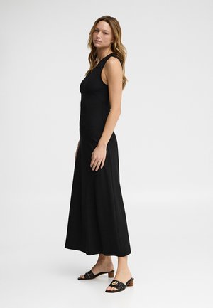 Kobieta o długich włosach, ubrana w czarną, bezrękawnikową suknię maxi i czarne sandały na obcasie, stojąca bokiem na białym tle.