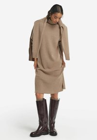 Beiges Strickkleid mit Rollkragen und gerippter Textur; kombiniert mit hohen, glänzenden burgunderfarbenen Stiefeln mit Strickoberteil.
