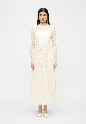 Mujer con cabello largo y oscuro, vestida con un vestido blanco de manga larga de encaje con patrones florales y zapatos blancos puntiagudos, de pie frente a un fondo liso.