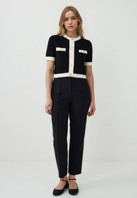Cardigan corto nero a maniche corte con bordi crema, bottoni dorati e due tasche frontali, abbinato a pantaloni neri su misura e ballerine nere.
