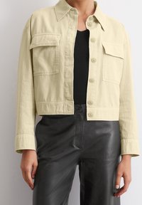 Light yellow cropped jacket with a button-up front, two chest pockets, and a collar, paired with black leather pants. Smooth texture.