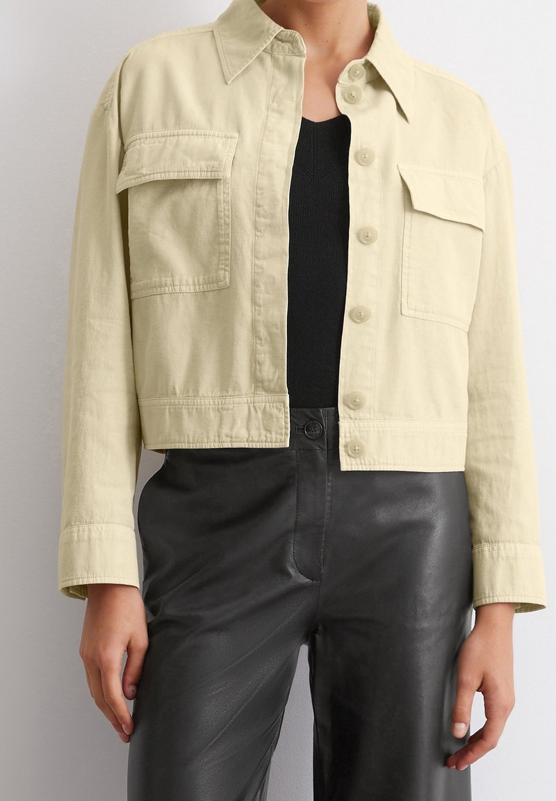Light yellow cropped jacket with a button-up front, two chest pockets, and a collar, paired with black leather pants. Smooth texture.