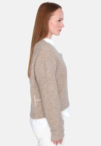 Beige ullgenser med kort design, ribbestrikket hals og lange ermer, med en hvit detalj på siden.