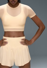 Woman wearing a beige short-sleeve crop top and loose high-waisted shorts standing with hands on hips against a plain gray background.