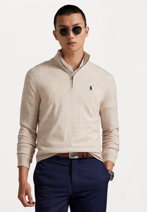 Man wearing beige quarter-zip sweater with navy embroidered logo, white shirt, navy pants, brown belt, watch, and round sunglasses standing against white background.
