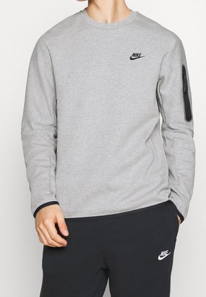 Mężczyzna ubrany w jasnoszarą bluzę Nike z kieszenią na zamek na rękawie i czarne spodnie dresowe Nike z logo na udzie.