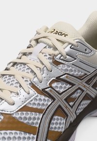 Nærbilde av hvit og sølvfarget Asics løpesko med beige lisser og brune detaljer, som viser teksturert mesh og logodetaljer.