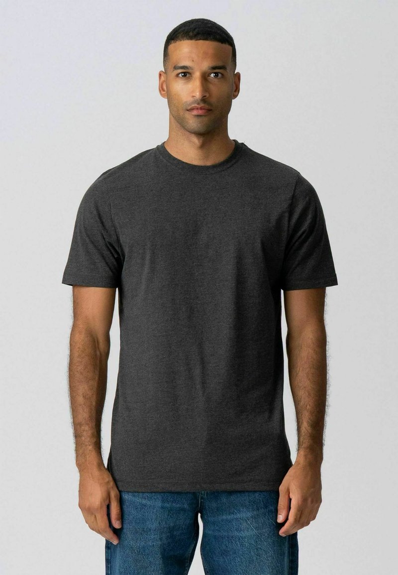 Homme aux cheveux courts portant un t-shirt gris foncé à col rond et un jean bleu, debout devant un fond uni gris clair.