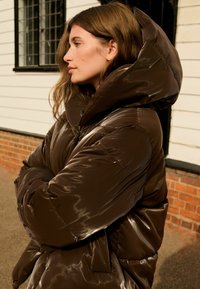 Hnědá lesklá péřová bunda s vysokým límcem a oversize kapucí. Obsahuje prošité panely a řasený pas pro tvarování.