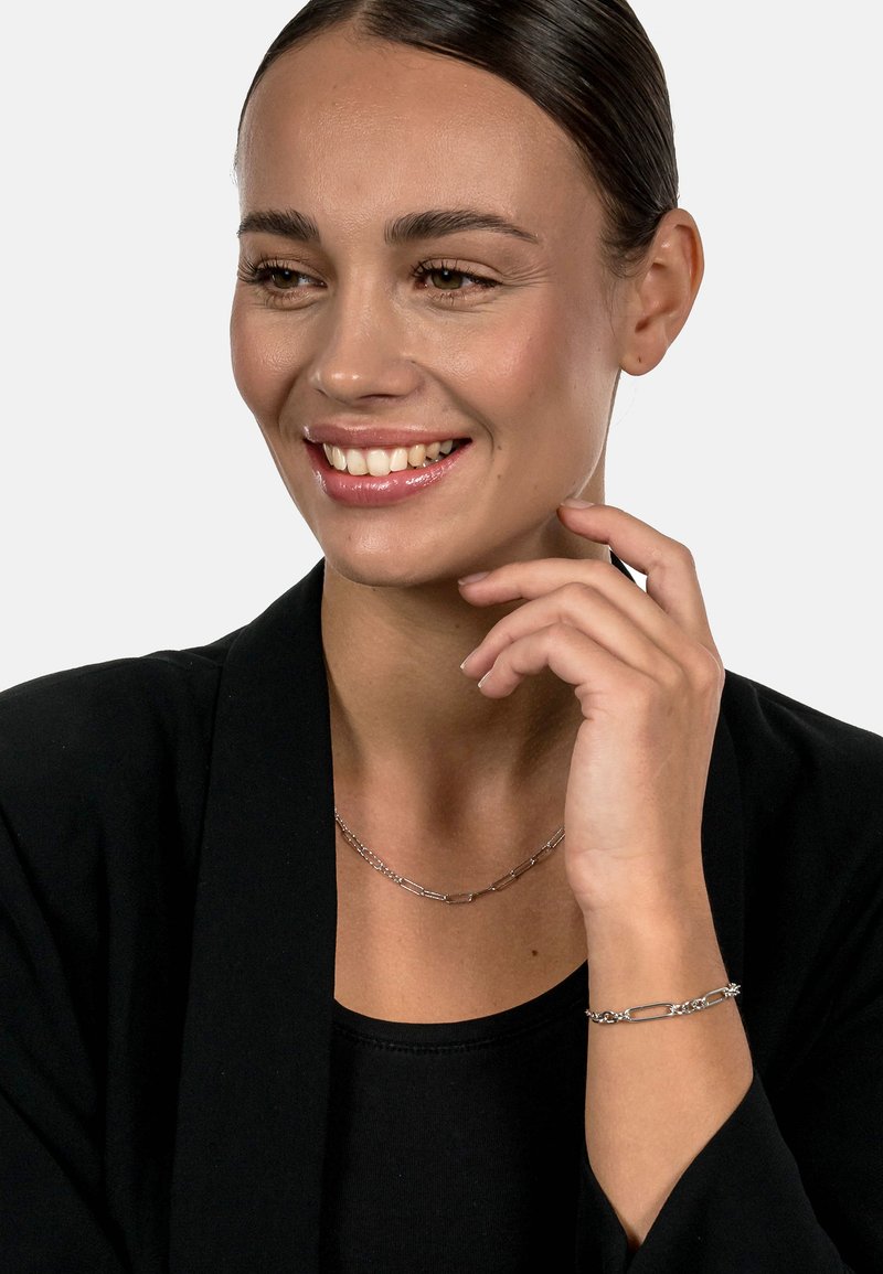 Silver link necklace and bracelet on a person, featuring elongated rectangular links with a glossy finish. Black top and blazer visible.