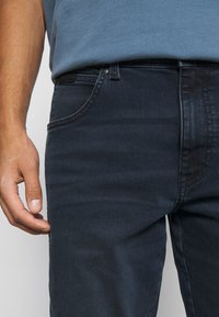 Jeans de mezclilla azul oscuro con un corte ajustado, que presentan texturas sutiles, bolsillos frontales y costuras en contraste, mostrados con una mano descansando en el lado.
