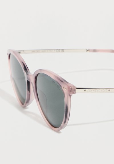 Στρογγυλά γυαλιά ηλίου με διαφανή ροζ πλαίσια και σκουρούς φακούς, με χρυσά μεταλλικά μπράτσα χαραγμένα με το λογότυπο "Jimmy Choo".