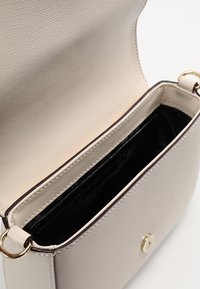 Beige käsilaukku, jossa on teksturoitu ulkopinta, avoin läppä ja musta sisusta. Varustettu yksinkertaisella kultaisella lukolla ja yhdellä sisäosastolla.