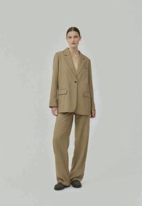 Abito beige su misura con giacca a un bottone, revers a punta e due tasche frontali, abbinato a pantaloni larghi. Texture liscia, vestibilità moderna.