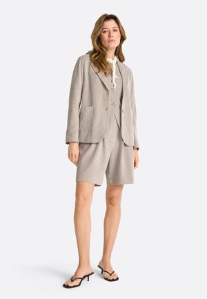 Frau steht und trägt ein beigefarbenes, mit Nadelstreifen versehenes Sakko, passende Shorts mit hoher Taille, eine weiße Bluse mit Schlips und schwarze Sandalen mit Absatz.