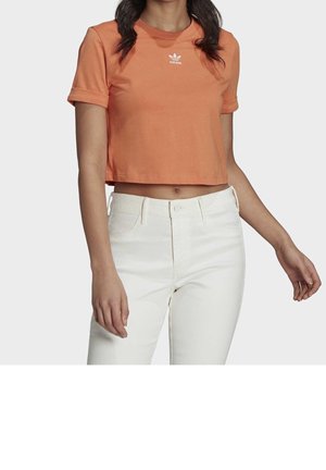 Kobieta w pomarańczowym, krótkim t-shircie Adidas i białych spodniach z wysokim stanem, stojąca z ramionami swobodnie opuszczonymi wzdłuż ciała.