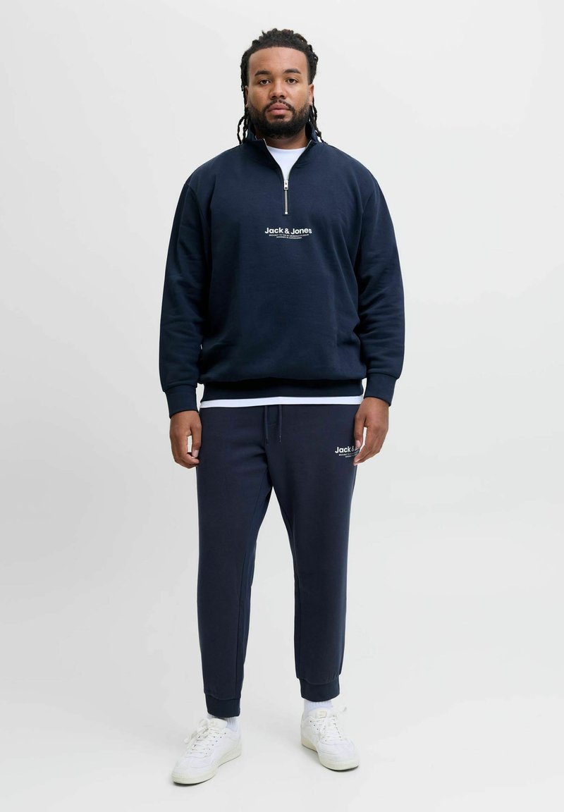 Marinblå zip-up sweatshirt med "Jack & Jones"-logotyp, som matchas med slim fit sweatpants. Vita sneakers kompletterar outfiten.