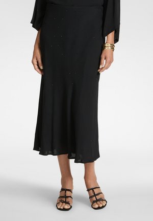 Frau trägt einen schwarzen Midirock mit dezentem Glitzern, goldene Armreifen und schwarze Riemchen-Sandalen mit offenem Zehenbereich vor weißem Hintergrund.