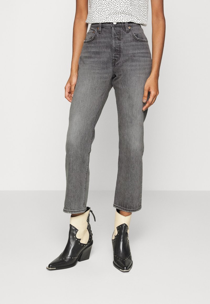 Femme portant un jean gris taille haute et des bottines à bout pointu noires et blanches avec des détails cloutés sur un fond clair.