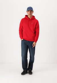 Felpa rossa con tasca frontale e cordino, abbinata a jeans blu scuro e scarpe nere. Il modello indossa un cappellino blu.