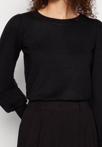 Maglione nero a maniche lunghe in maglia con scollo rotondo e spalle leggermente imbottite, indossato sopra pantaloni neri a vita alta e sartoriali.