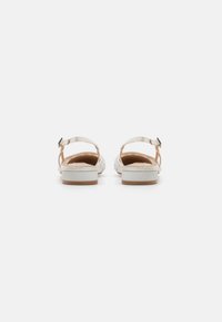 White open-toe sandals with ankle straps, textured upper, and a low, solid brown heel. Soft interior lining for comfort.
