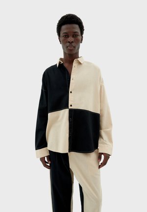 Button-up shirt featuring a two-tone design in black and beige, with contrasting stitching and long sleeves. Made from soft fabric.