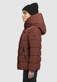 Rote Pufferjacke mit großer Kapuze, genähten Abschnitten und einer seitlichen Reißverschlusstasche. Der Stoff ist glatt und hat ein warmes, isoliertes Aussehen.