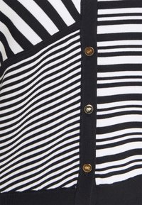 Zwart-wit gestreepte cardigan met verschillende streep patronen, gebreide textuur en gouden knopen. Het ontwerp bevat effen zwarte accenten.