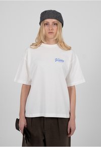 Valge lühikeste varrukatega puuvillane t-särk, millel on vasakul rinnal väike sinine „Keelatud“ graafika. Sobitage pruunide kõrge vöökohaga pükstega.