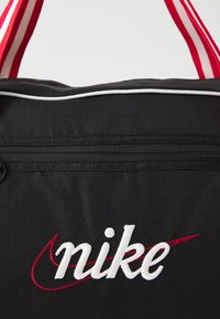 Black fabric bag featuring white and red "nike" logo, red and white striped straps, and a smooth zipper pocket on the front.