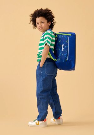 Blue backpack with geometric patterns, worn over a green and white striped shirt. The child wears dark blue pants and white sneakers.