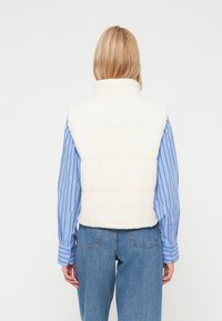 Bela, puhasta brezrokavnik z visokim ovratnikom, ki ima prešito teksturo, nosi se čez modro črtasto srajco in je v kombinaciji s svetlo modrimi kavbojkami.