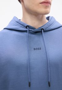 Primo piano di una persona che indossa un felpa blu con cappuccio e cordini, con il logo "BOSS" centrato sul petto, vista parziale della mascella e del collo.