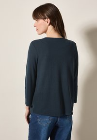 Tumma teal-värinen neule, jossa on pitkät hihat ja pyöreä pääntie. Hieno tekstuuri ja rento malli. Yhdistetty sinisiin farkkuihin.