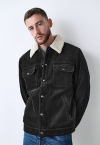 Chaqueta de pana negra con un collar de piel sintética color crema, frente de botones, dos bolsillos en el pecho y textura acanalada detallada. Llevada con jeans de mezclilla.