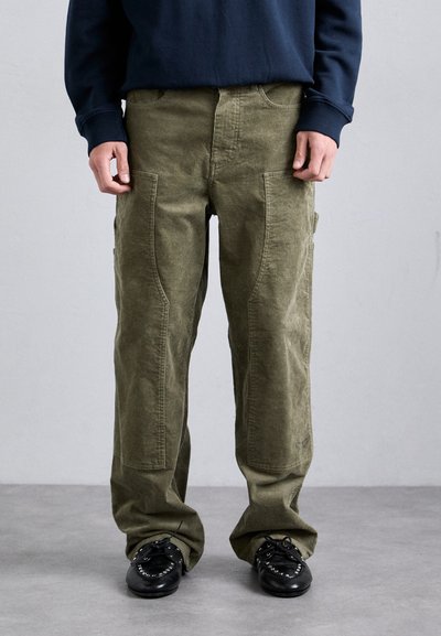 Person in olivgrünen Cordhosen mit großen vorderen Taschen, navyblauem Langarmoberteil und schwarzen Schnürschuhen steht auf einem grauen Boden.