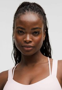 Light pink tank top with thin straps, featuring a smooth texture. The model has braided hair and a neutral expression, showcasing facial features.