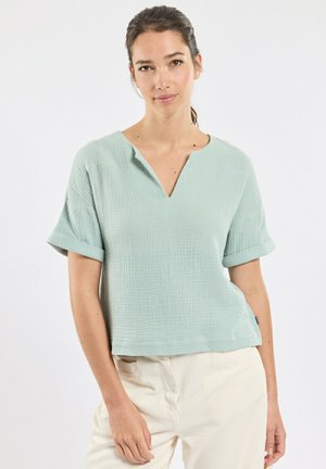 Femme aux cheveux foncés attachés, portant un haut texturé à manches courtes vert clair et un pantalon beige clair, debout devant un fond blanc.